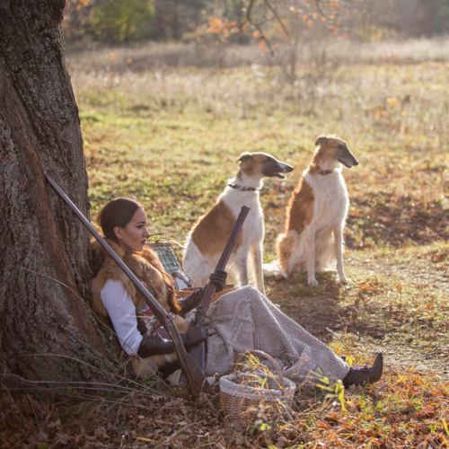 A woman in vintage clothing sits against a tree holding a rifle, with two Borzoi dogs sitting nearby in a sunlit, grassy field. A woven basket rests on the ground beside her.