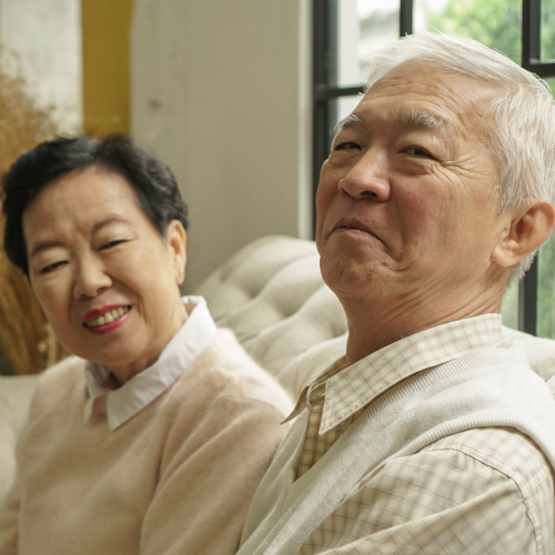 An older man and woman sit together on a sofa, smiling warmly. The man, wearing a light shirt and vest, adjusts his hearing aids while the woman in a light sweater looks on. Soft daylight comes through a window behind them.