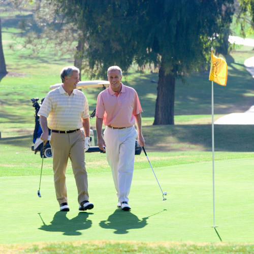 Two older men, smiling and holding golf clubs, walk together on a sunny golf course near a hole with a yellow flag. Both wear discreet hearing aids, enjoying clear conversation as a golf cart and green trees fill the background.