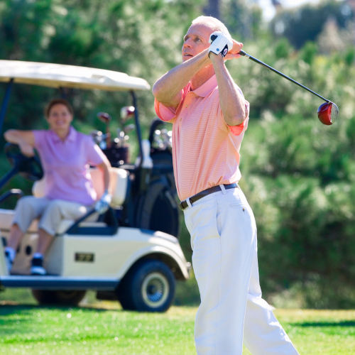 An older man in a pink shirt and white pants swings a golf club on a course, while a woman in a pink top sits in a golf cart nearby, the lush greenery creating a serene backdrop—much like the calm precision of tympanometry or otoscopy.
