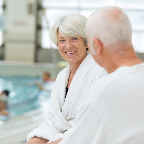 Two older adults in white bathrobes sit by an indoor swimming pool, smiling and talking. The background shows other people swimming and bright, natural light coming through large windows.