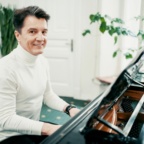 A man in a white turtleneck sweater sits at a grand piano, smiling at the camera in a bright, elegant room with green plants and a white door in the background.