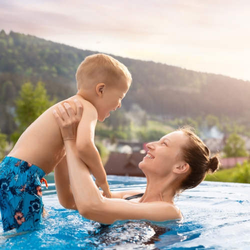 A woman in a pool lifts a smiling young boy wearing blue swim trunks. Outdoors with trees and hills in the background, the sun shines brightly—creating a joyful scene reminiscent of the positive spirit often seen after successful audiometry tests.