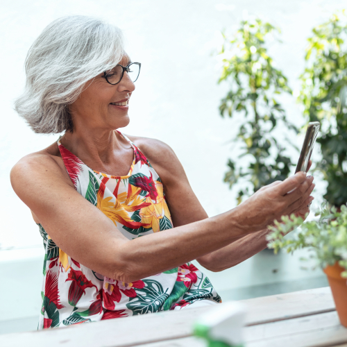 Smiling older woman with gray hair, glasses, and discreet hearing aids wears a colorful floral dress as she takes a selfie with her smartphone while sitting at an outdoor table surrounded by plants.