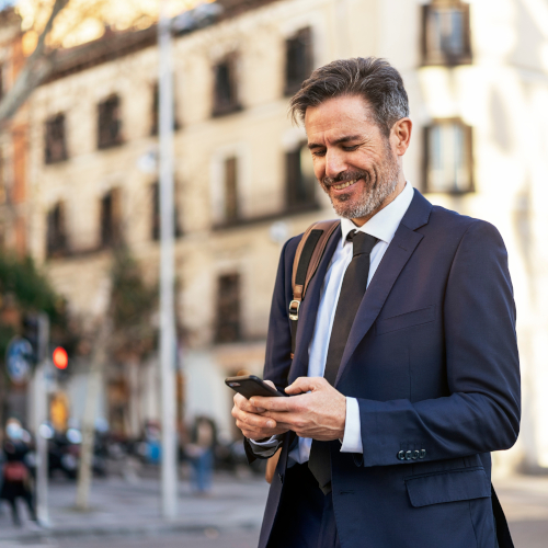 A man in a dark suit and tie, wearing hearing aids, smiles while using his smartphone outdoors in a city. He has a beard, carries a shoulder bag, and stands before a building with balconies and blurred street activity behind him.