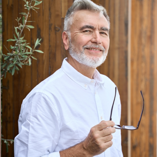 An older man with gray hair and a beard smiles while holding eyeglasses in one hand. He is wearing a white button-up shirt and standing outdoors in front of a wooden wall with some greenery beside him.