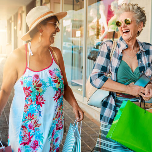 Two older women laugh joyfully while carrying shopping bags on a sunny day. One wears a floral dress and sunhat, the other a plaid shirt and sunglasses. They walk together outside shops, enjoying each others company.