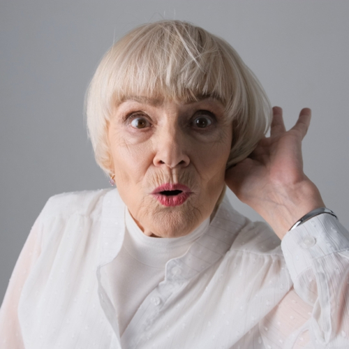 Elderly woman with short blonde hair wearing a white blouse, holding her hand to her ear and looking surprised, as if trying to hear something better, possibly considering hearing loss treatment.