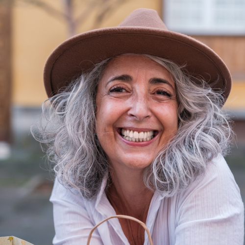 Smiling woman with wavy gray hair wearing hearing aids, a brown hat, and white shirt, sitting outdoors. The background is softly blurred, featuring a yellow building and a window.