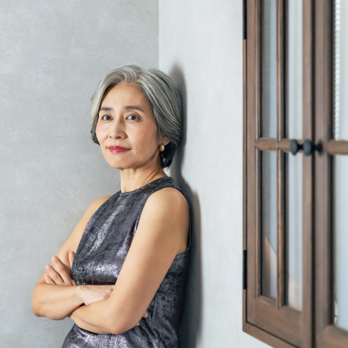 An older woman with short gray hair, wearing discreet hearing aids and a sleeveless metallic top, stands indoors with her arms crossed, leaning against a light-colored wall near a wooden-framed window, smiling softly.