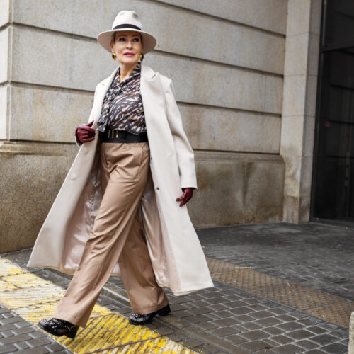 A stylish older woman walks confidently on a city sidewalk, wearing a wide-brim hat, cream long coat, patterned blouse, tan trousers, maroon gloves, and black shoes. The background features a stone building.