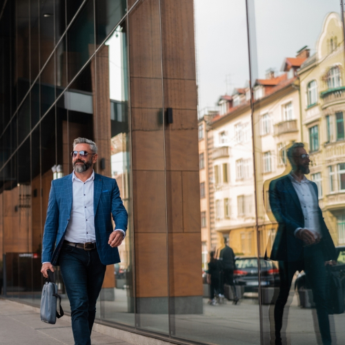 A man in a blue blazer and sunglasses walks confidently on a city sidewalk, holding a gray bag. Buildings and his reflection are visible in the glass windows next to him.