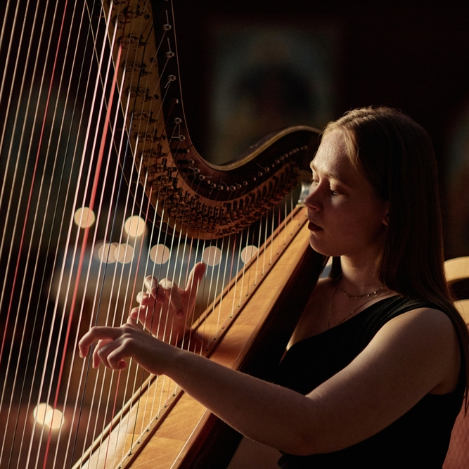 A woman in a black dress plays a harp, her eyes closed in concentration, with warm light illuminating her and creating a soft, blurred background with bokeh effects.