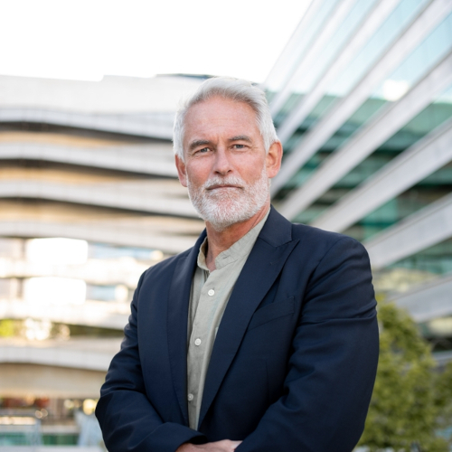A mature man with gray hair and a beard, wearing a dark blazer and light shirt, stands confidently outdoors in front of modern glass and concrete buildings, embodying the assurance that comes with effective hearing loss treatment.