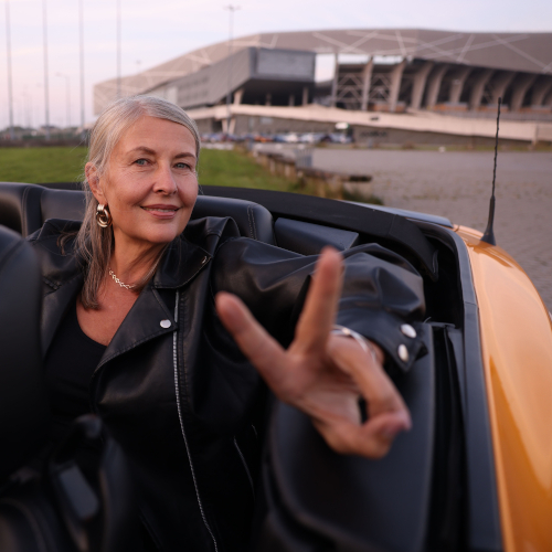 An older woman with gray hair, wearing a black leather jacket, sits in a yellow convertible and smiles while making a peace sign, with a modern stadium visible in the background.
