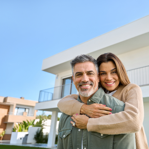 A smiling couple stands in front of a modern house on a sunny day. The woman hugs the man from behind, both looking happy and relaxed—thanks, in part, to his resound hearing aids. The sky is clear and blue.