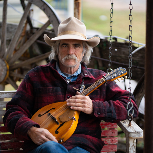An older man with a gray mustache, wearing a cowboy hat and plaid jacket, sits on a porch swing holding a mandolin. A rustic wooden wagon wheel is visible in the background.