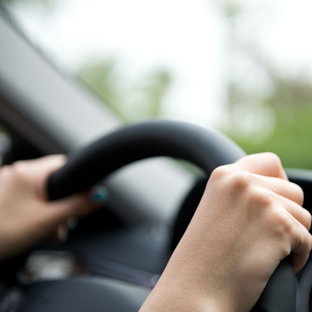 Close-up of a person’s hands gripping a black steering wheel inside a car, with a blurred green background visible through the window.
