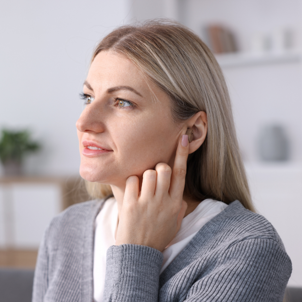 A woman with blonde hair touches her ear while looking to the side. She is wearing a gray sweater and sitting indoors in a softly lit room.